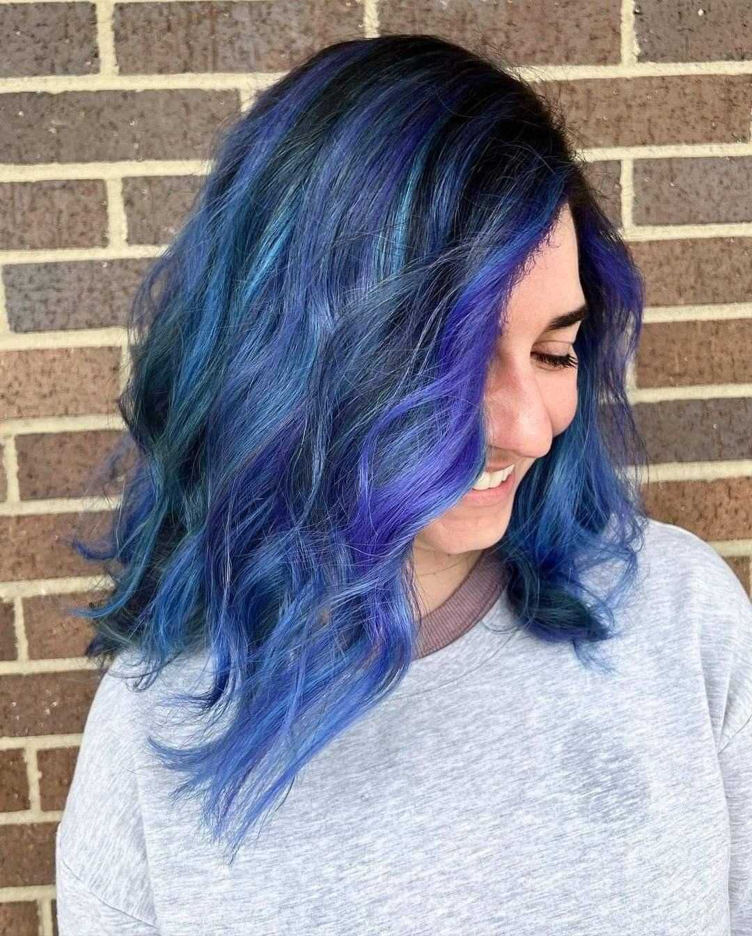 Woman with wavy, vibrant blue and purple hair smiling against a brick wall.