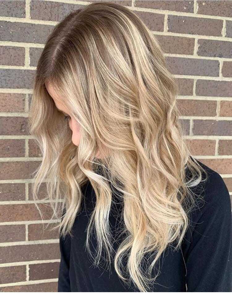 Woman with long, wavy, blonde hair stands against a brick wall looking down.